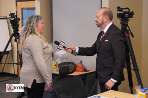 A reporter interviews a woman in an office setting. The room has a table with papers, a bowl of oranges, and a camera set up on a tripod.