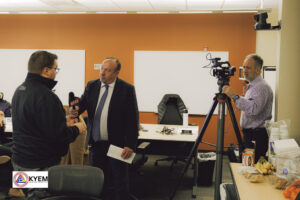 A reporter interviews a man in an office setting. A cameraman films the interview with a camera set up on a tripod. 