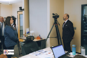 A reporter interviews a woman in an office setting. The room has a table with papers, a bowl of oranges, and a camera set up on a tripod.