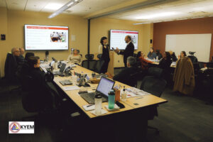 People in a conference room attend a training; most of the people are seated at tables, some with laptops. Two people stand in the front of the class, one interviewing the other. Presentation slides are displayed on a screen at the front of the room.